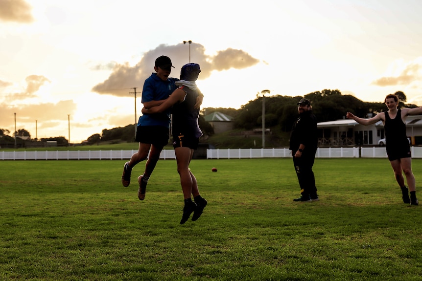 Two kids jump in the air in a hug will two others look on across a country football ground