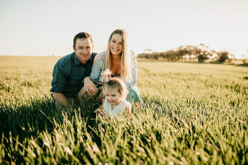 Rebecca kneeling in a field with her husband and young daughter.