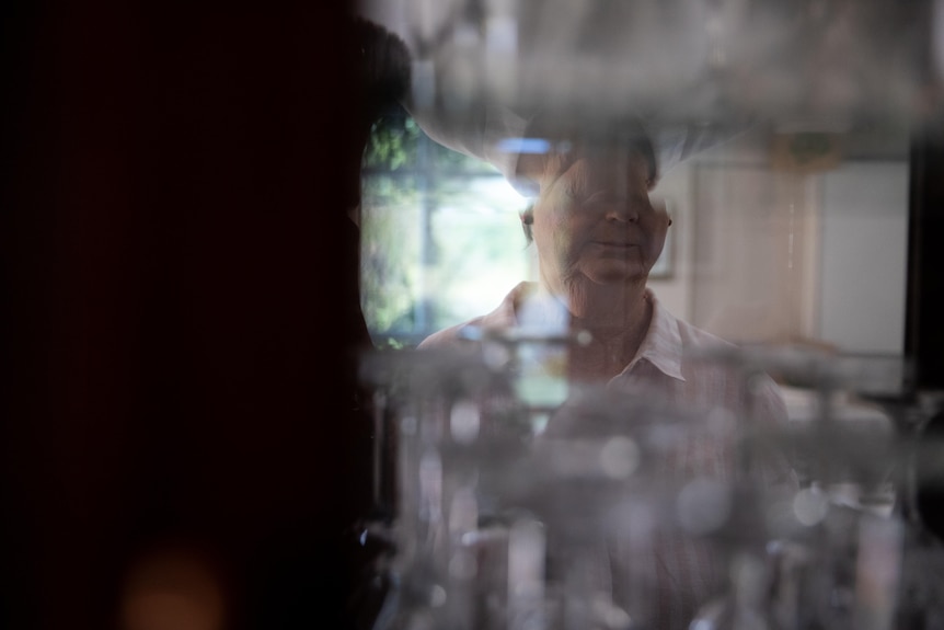 A woman with a solemn expression looks into the mirror of a glass cabinet, her eyes are not visible.