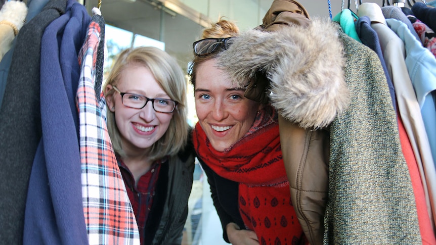 Two young girls peak through a rack of coats, smiling.