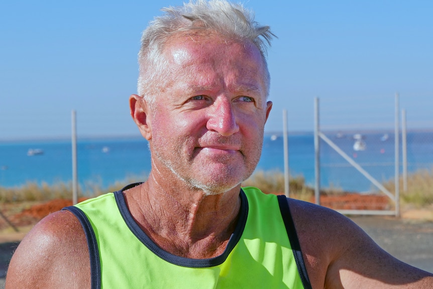 A middle-aged man in a brightly-coloured singlet stands in front of the ocean.