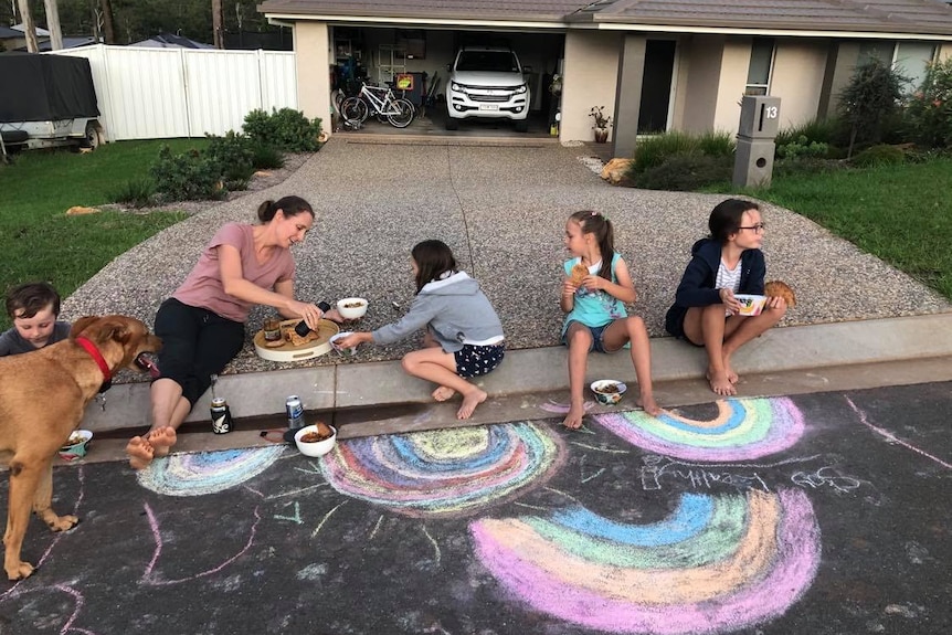 A woman and children sit on a driveway eating a meal, with chalk drawings on the ground.