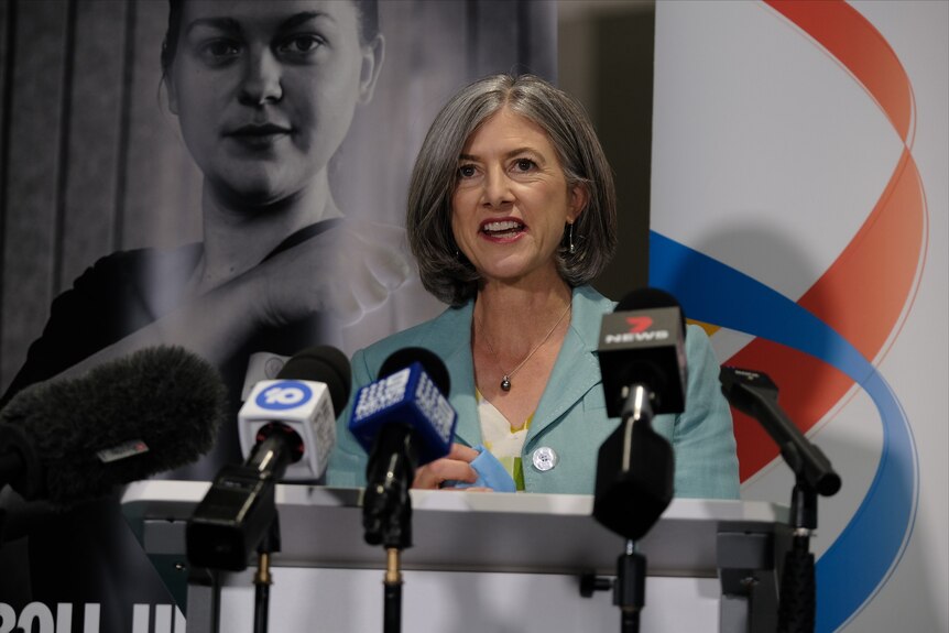 A woman wearing a light blue jacket stands in front of a picture of a woman and behind microphones