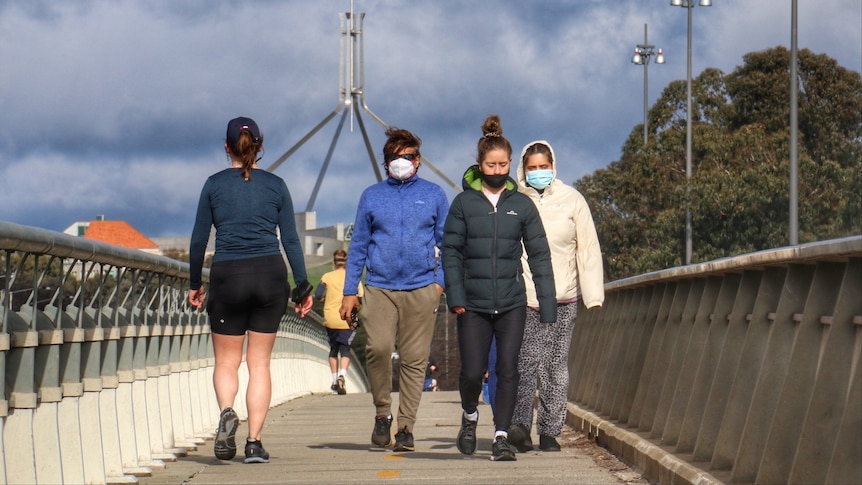 People in face masks walk over a bridge with parliament house in the background.
