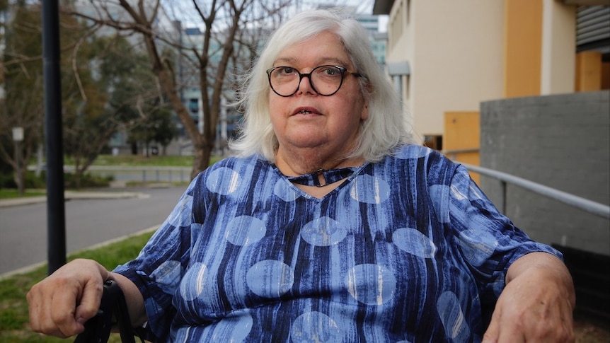 A woman in glasses and a blue patterned shirt sits in front of a road and apartment block