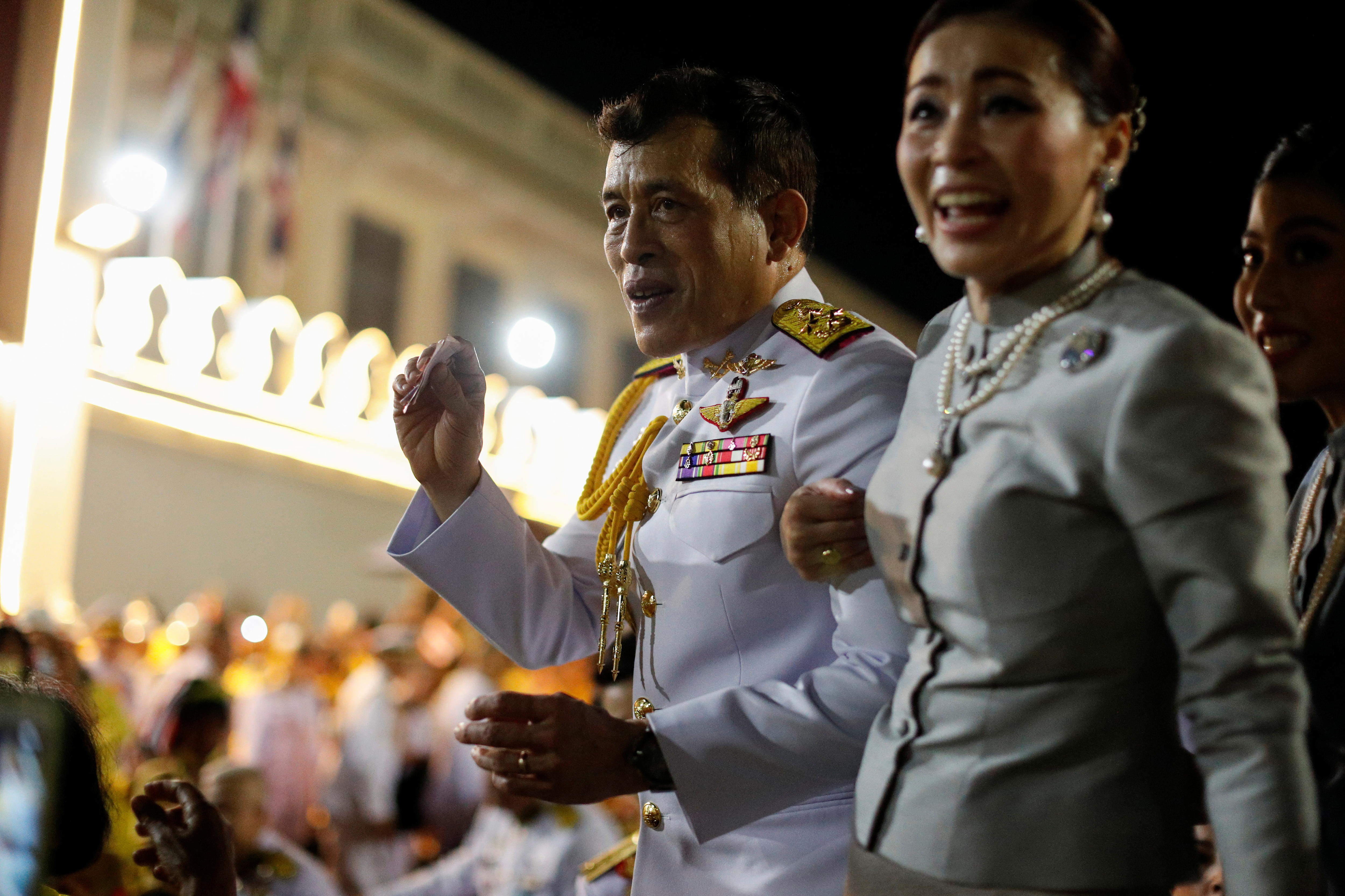 A middle-aged Thai man in a white uniform, and a woman in a light-coloured dress, smile and wave to supporters outside at night.