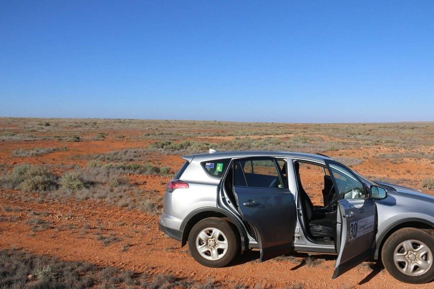 Car in the foreground with red dust and desert in the background and clear blue sky.