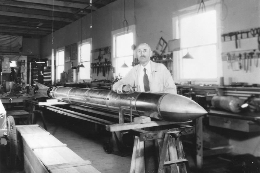 Man in suit and lab coat rests arm on a rocket inside a workshop.