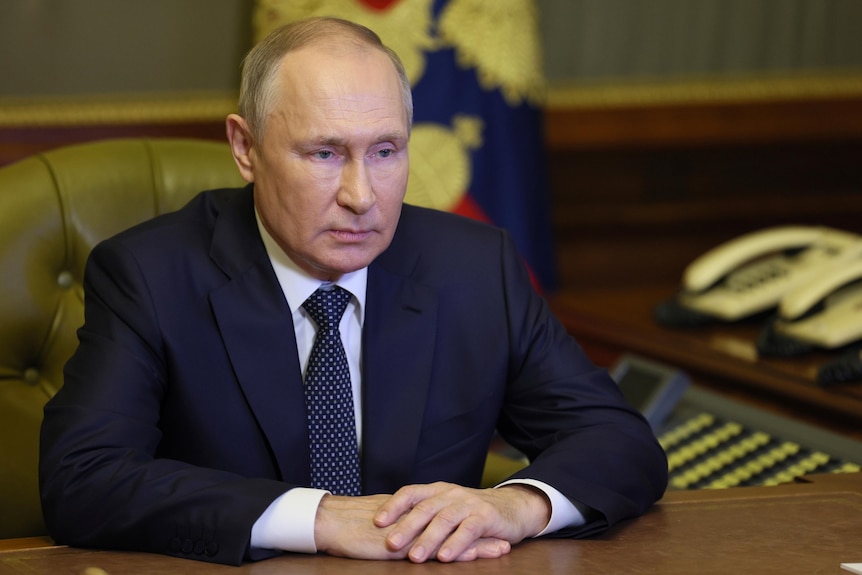 Russian President Vladimir Putin sits in a chair with his hands on a desk. 