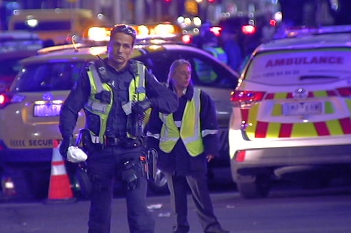 A police officer in uniform walks on the street with an ambulance in the background.