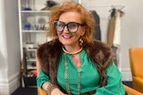 A woman with red hair and glasses in front of a clothes and make-up stand 