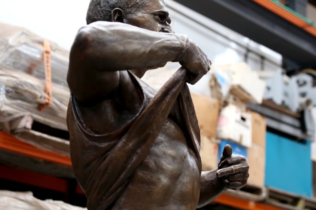 A bronze statue of a football player pulling up his top and pointing to his belly sits in a workshop.
