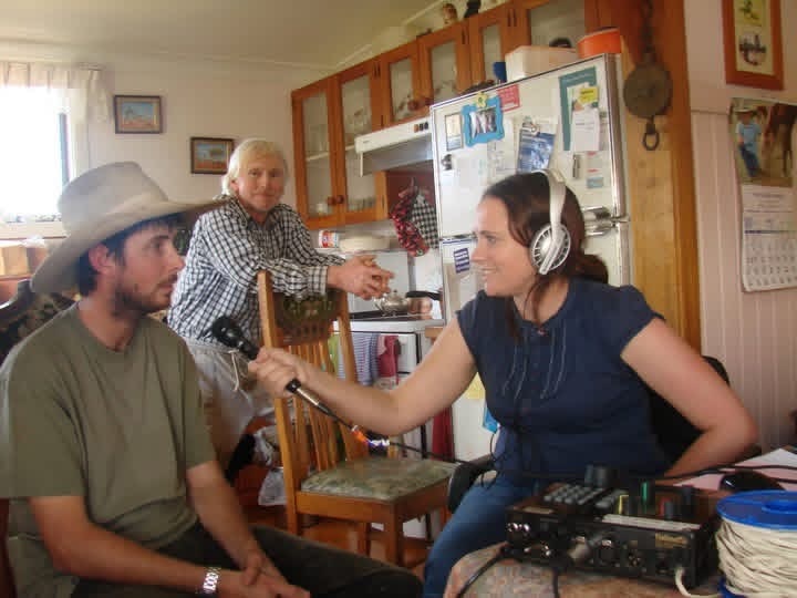 a woman, right with headphones and holding a microphone talks to a man with a bush hat in a kitchen