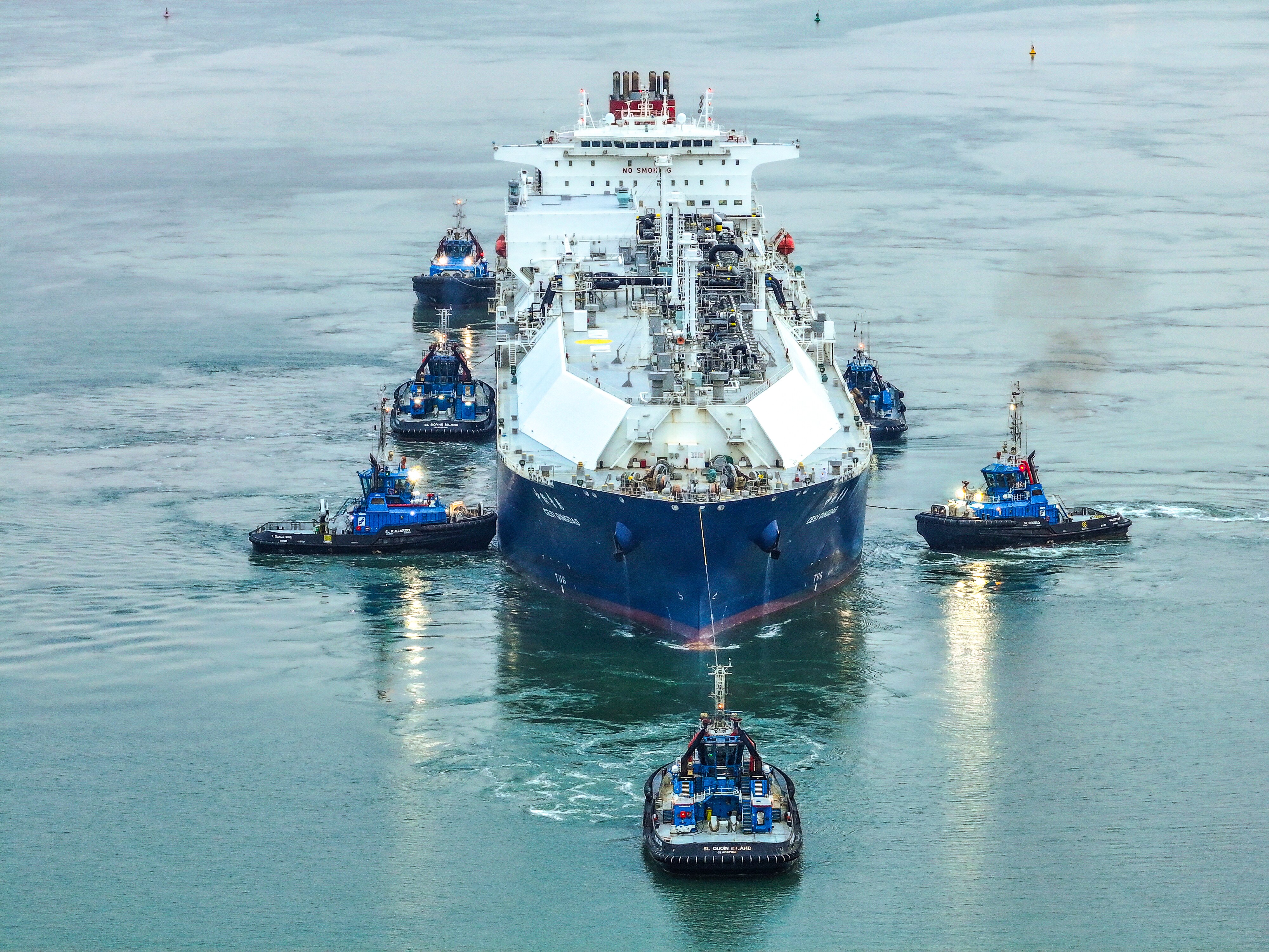 Front-on view of large vessel being towed by several tug boats through water