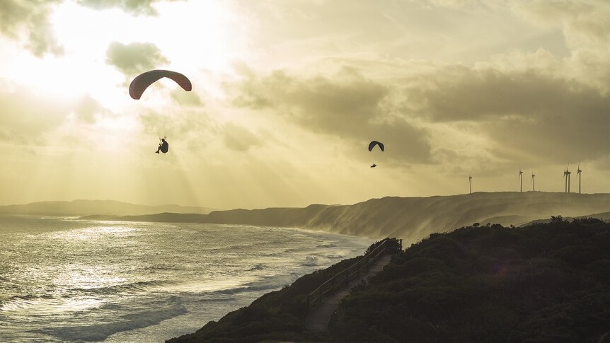 The sun sets behind two paragliders flying over the coast, with wind turbines on the coast in the distance.
