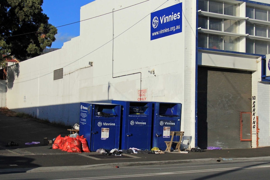 Three blue charity bins with bags and bits and pieces lying around them