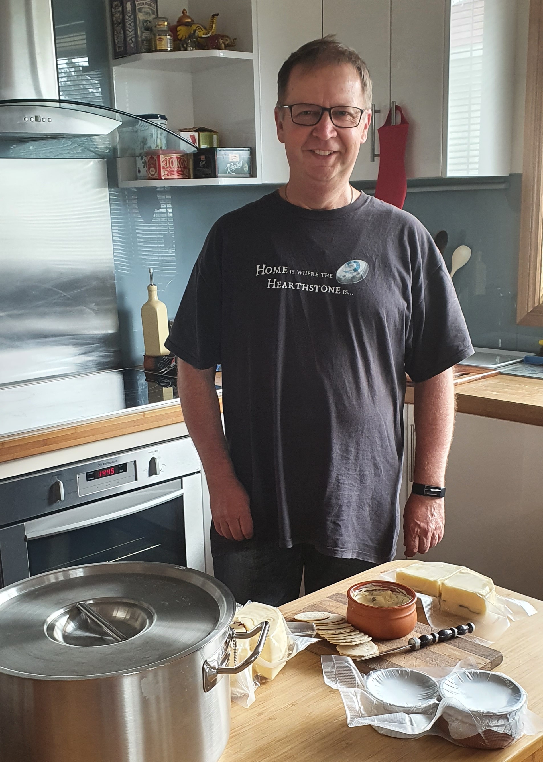 Man wearing glasses and a black t-shirt stands in his kitchen next to a bench with homemade cheeses.