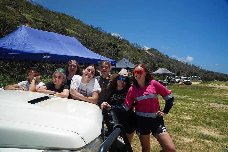 Seven girls smile, leaning on a car at the beach.
