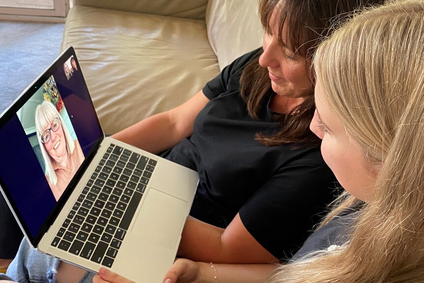 Two women sit on a couch with a laptop chatting on Facetime with their relative