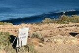A white sign reading KEEP OUT OF WATER sits on top of a rocky cliff overlooking the deep blue ocean in the background. 