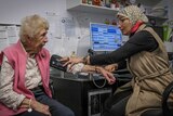 A woman wearing a patterned hijab take the blood pressure of an elderly woman wearing a pink vest.