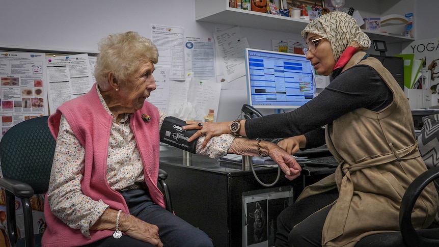 A woman wearing a patterned hijab take the blood pressure of an elderly woman wearing a pink vest.