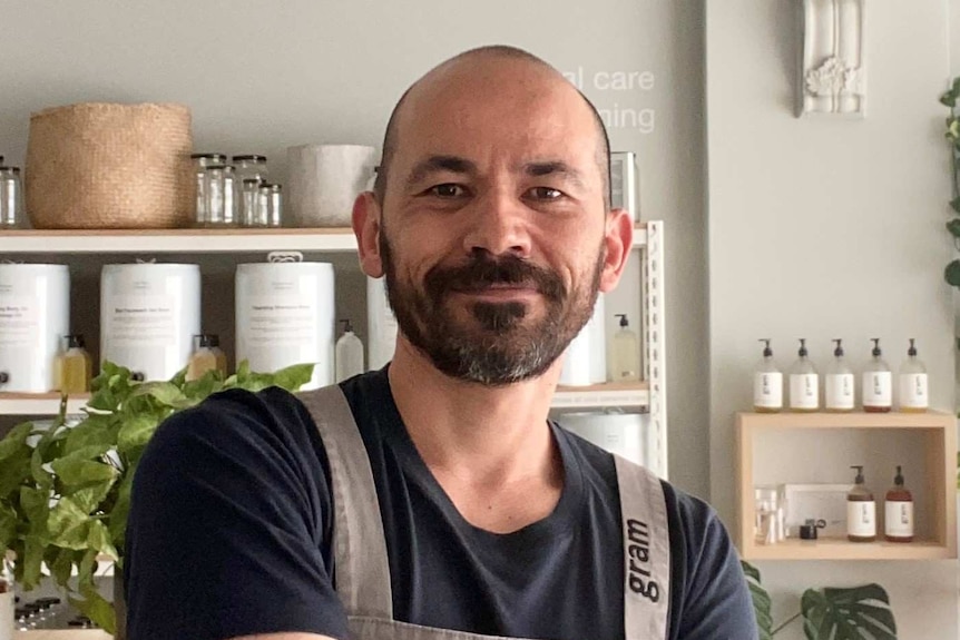 A man with a bald head, beard and moustache stands in front of shelves with his arms crossed in front of him.