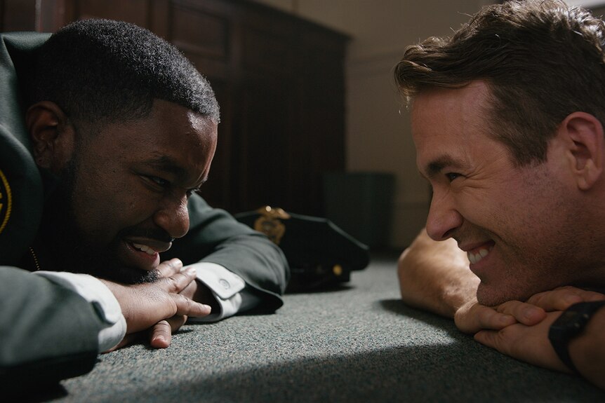 Film still from Free Guy (2021) showing Lil Rey Howery and Ryan Reynolds lying down and grinning at each other.