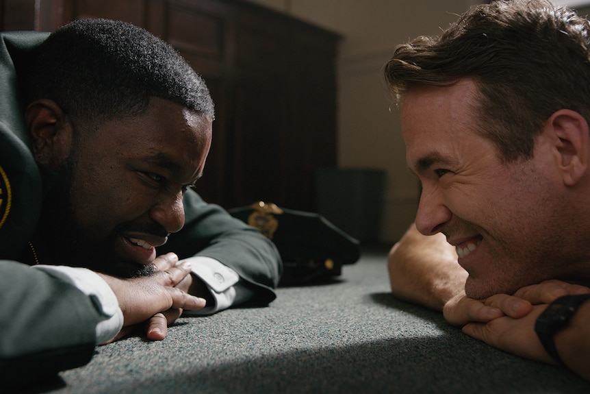 Film still from Free Guy (2021) showing Lil Rey Howery and Ryan Reynolds lying down and grinning at each other.