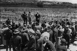 a large crowd of men in suits and bowler hats watch on as a man gives a speech at a set of saleyards in 1925