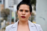 A close up shot of a woman with black hair, a mauve jacket and red lipstick.