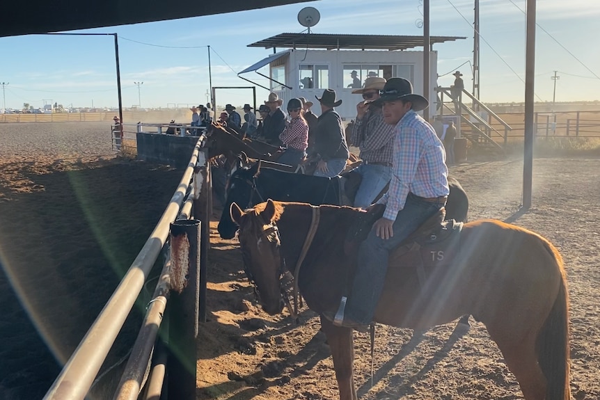 Cowboys and girls wearing hats, boots, jeans and collared shirts line up at a fence in a dusty arena, there's sun flares.