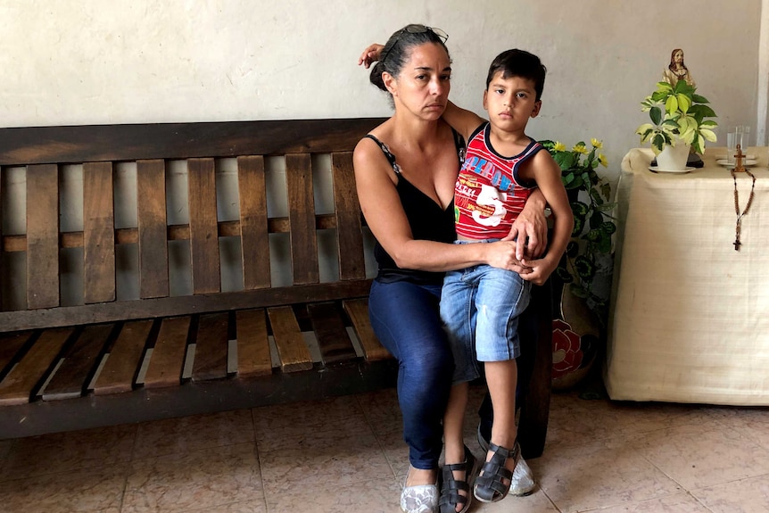 A bereaved mother sits on a wooden bench with a child and a statue of Jesus on the table next to her.