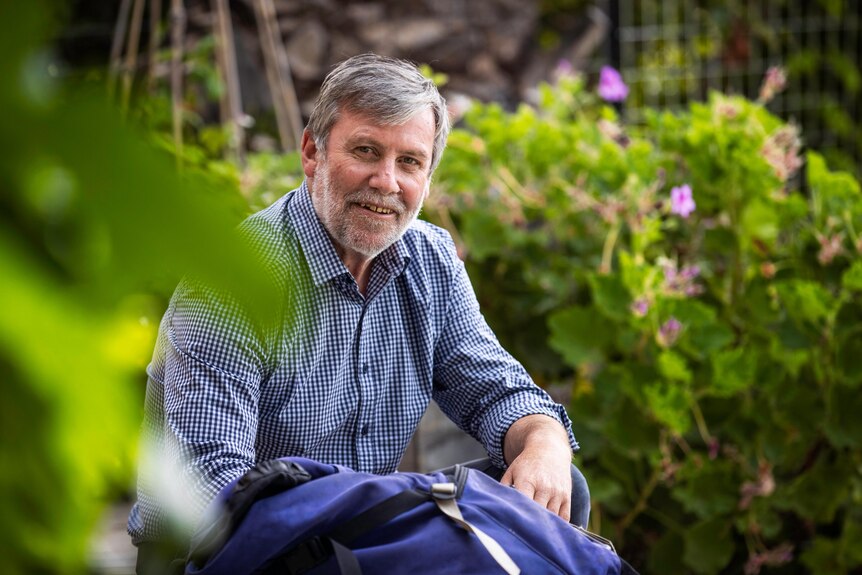 A man sits among out of focus greenery in a garden, with the top of a bushwalking pack visible next to him