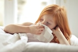A young girl with red hair tucked into bed, lying on her side and blowing her nose.