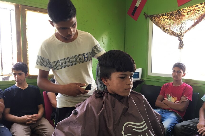 16-year-old Arabi practices his barbering skills on a young boy, while others wait for their turn.