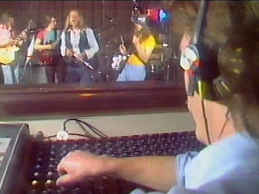 A recording engineer operates a recording panel while, through a window, a band is visible playing in a studio.