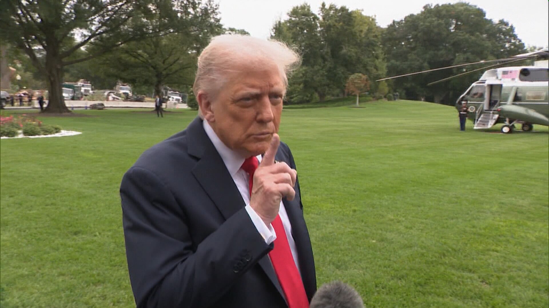 Donald Trump standing on grass at the White House holding his right index finger near his mouth.