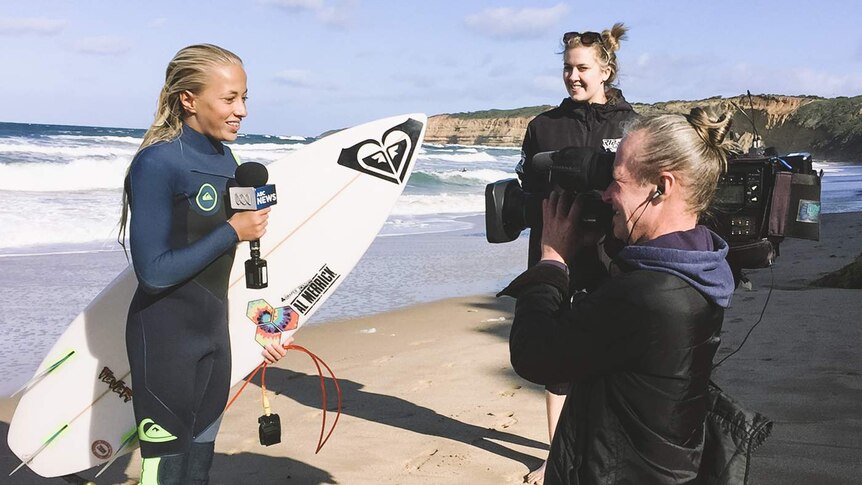 A girl on the beach holds her surfboard and an ABC News microphone while speaking to camera.