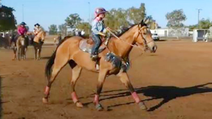 A girl wearing a pink and purple helmet sits on a saddle and rides a horse on red dirt under a blue sky.