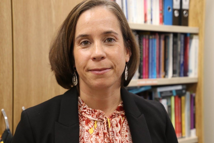 A close up of Katharina Wolf wearing a black jacket, standing in front of a book shelf looking into the camera.