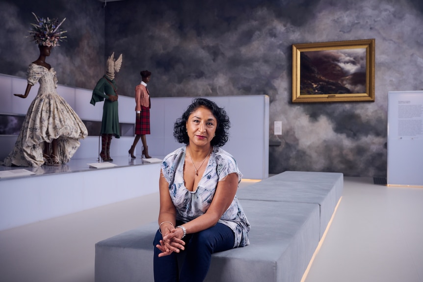 Middle-aged woman of colour with short dark curls wears blue and white patterned shirt and navy pants while seated in gallery.