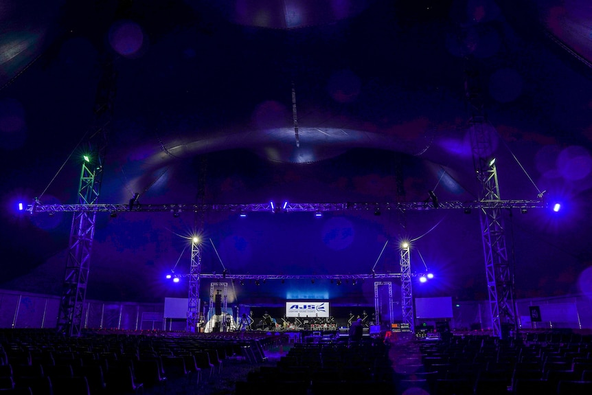 A blue glow emits from a large elevated stage inside a giant big top tent.