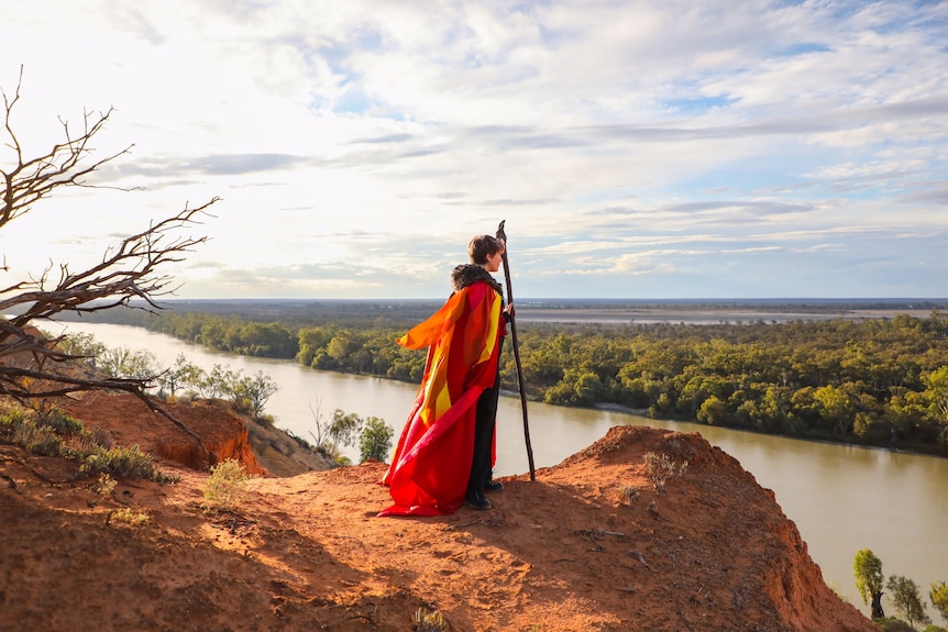 A person wearing a red and yellow and black cloak holds a stick and looks over the Murray River.