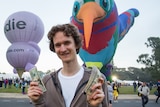 A young man stands holding money in both hands with large inflatable toys behind him at a park