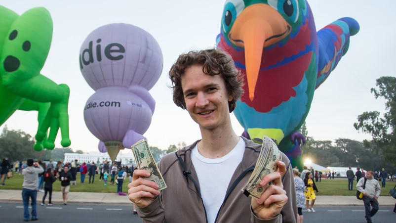 A young man stands holding money in both hands with large inflatable toys behind him at a park