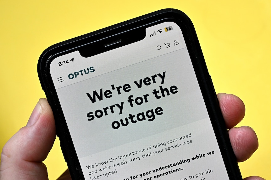 A close up of a hand holding a mobile phone, which is displaying an apology to customers written on the Optus website.