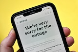A close up of a hand holding a mobile phone, which is displaying an apology to customers written on the Optus website.