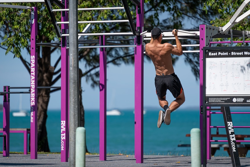 A photo of a muscly man doing chin-ups at an outdoor gym near the beach in Darwin. He is wearing black shorts. 