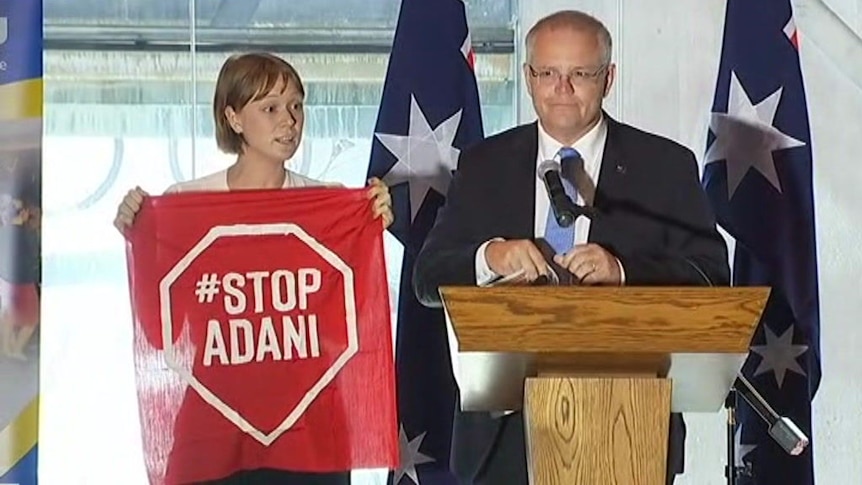 A Stop Adani protester standing next to Scott Morrison on the stage holding a Stop Adani sign.
